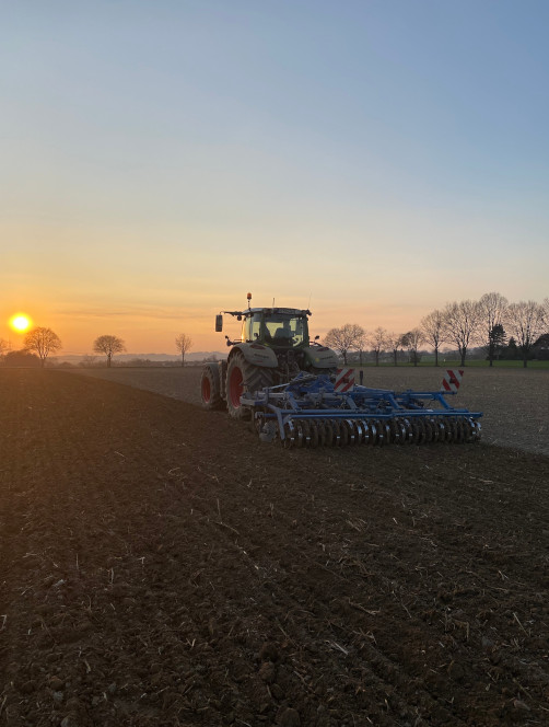 Ein Trecker bearbeitet ein Feld und fährt dabei dem Sonnenaufgang entgegen.